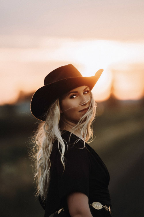 Vaaleahiuksinen nainen cowboy-hattu päässään. Taustalla näkyy kaunis auringonlasku.