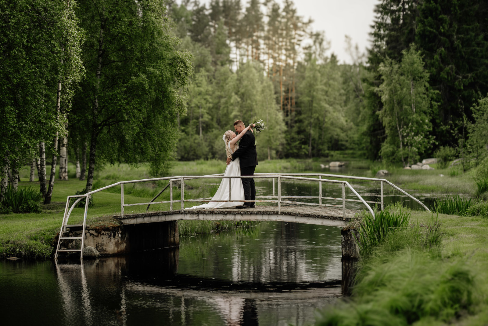 Hääpari halaa jokisillan päällä metsämiljöössä.