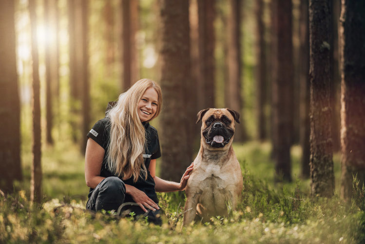 Bullmastiffi-koira ja omistajansa istuvat metsässä mäntyjen ympäröimänä.