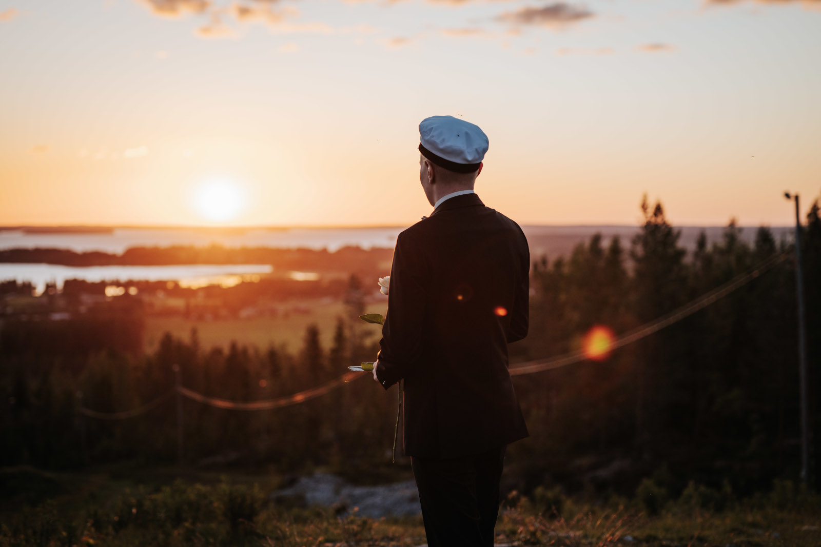 Tuore ylioppilas ruusu kädessä ja ylioppilaslakki päässä, kauniissa auringonlaskussa Lakeaharjun päällä.